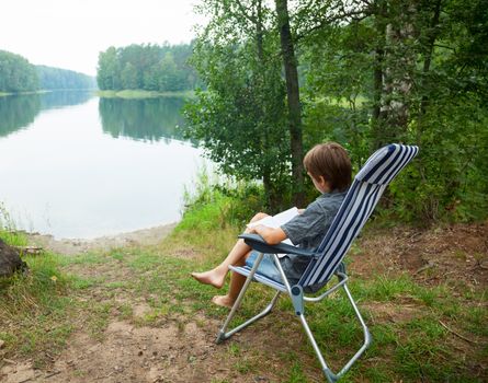 Boy sitting in deckchair reading a book on a lake shore