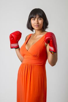 Young asian woman wearing a low cut orange dress, with her fist up wearing red boxing glove