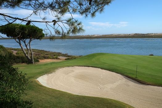 Overview of a Golf course and a sand trap, with pine trees and a stream of water behind