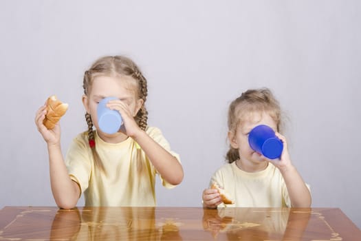 Two children sitting at the table, eating a muffin and drink of colored plastic cups