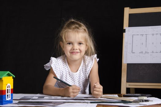 Four-year-old girl in the image of the architect. The girl sitting at the table with the drawings. In the background stands the Board with the drawing. Girl fun looking at the picture