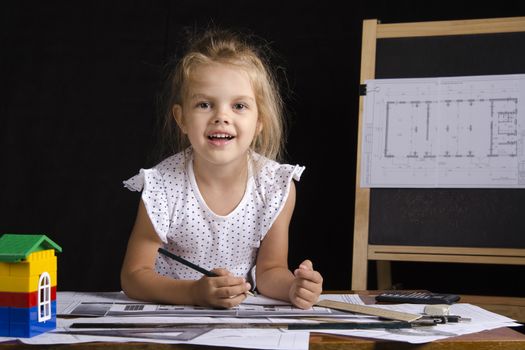 Four-year-old girl in the image of the architect. The girl sitting at the table with the drawings. In the background stands the Board with the drawing. Girl fun looking at the picture