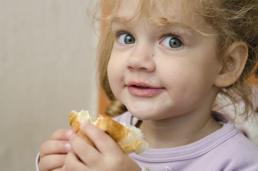 The little girl sits at a table and with enthusiasm eats a roll. Photo close up.