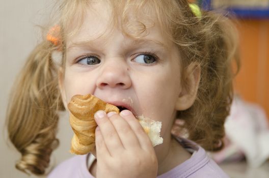 The little girl sits at a table and with enthusiasm eats a roll. Photo close up.