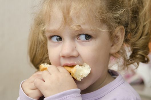 The little girl sits at a table and with enthusiasm eats a roll. Photo close up.