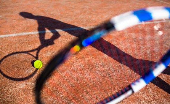 Shadow of a tennis player in action on a tennis court (conceptual image with a tennis ball lying on the court and the shadow of the player positioned in a way he seems to be playing it)