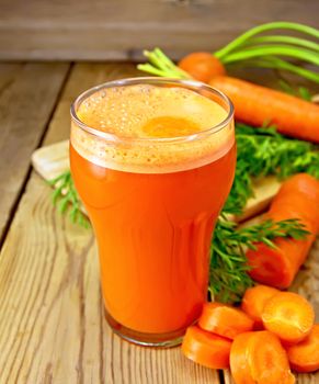 Carrot juice in a tall glass, carrots with greens on a wooden boards background