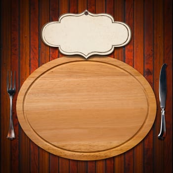 Wooden oval cutting board with empty label and silver cutlery, fork and knife on wooden table

