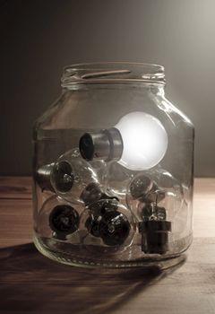 Illuminated light bulb inside a glass jar with several unlit lamps