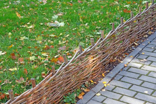 Braided twigs low fence separating diagonally lawn with fallen leaves and pavement of gray tiles                               