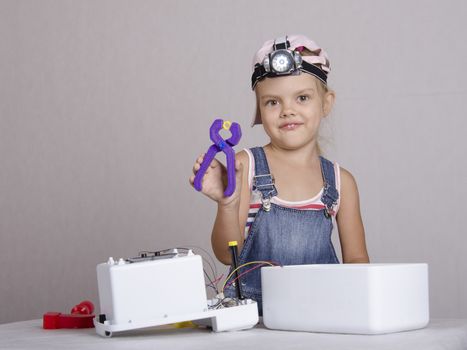 Girl playing in the repairer and repairs toy microwave. A girl Holds the pliers in which clamped nut and looks in a frame