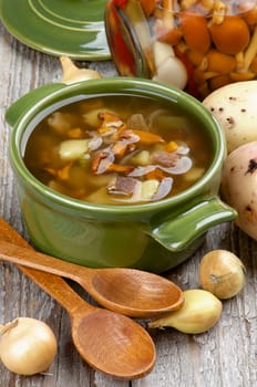 Delicious Vegetarian Soup with Chanterelle Mushrooms  in Green Pot with Raw Potato, Onion, Marinated Mushrooms and Wooden Spoons on closeup Rustic Wooden background