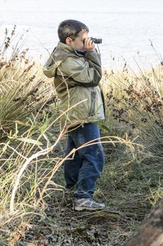 A school-aged boy looks through binoculars with a sea shore on the background.