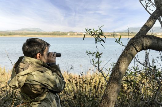 A school-aged boy looks through binoculars with a sea shore on the background.