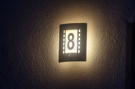 Illuminated house number, outdoor lamp with integrated number 8 at night on a white wall.