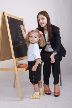  Two-year-old girl learns to paint a house on the blackboard chalk. Sitting beside educator who helps her. Girl and educator look in the frame
