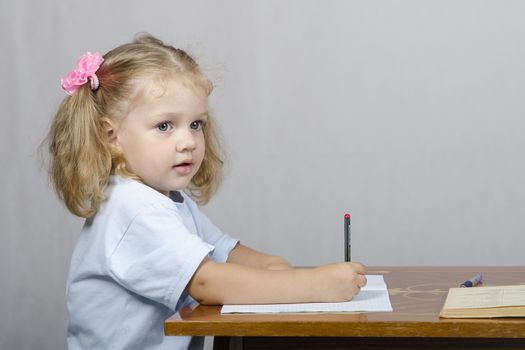 Little girl sitting at the table and writes handle in a notebook. The girl was distracted and looked to the right.