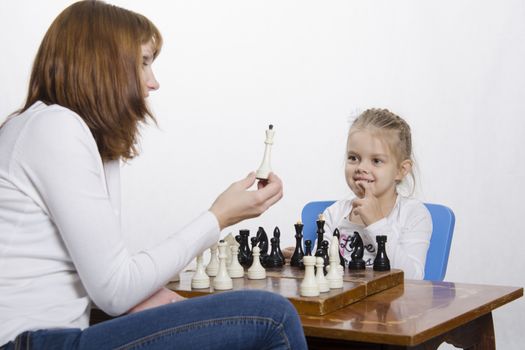 Mother and four-year-old daughter sitting at a table and play chess. Mom tells about the shapes