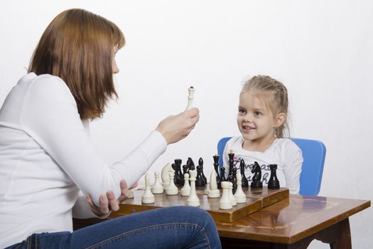 Mother and four-year-old daughter sitting at a table and play chess. Mom tells about the shapes