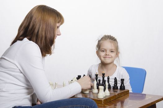 Mother and four-year-old daughter sitting at a table and play chess. Mom tells about the shapes