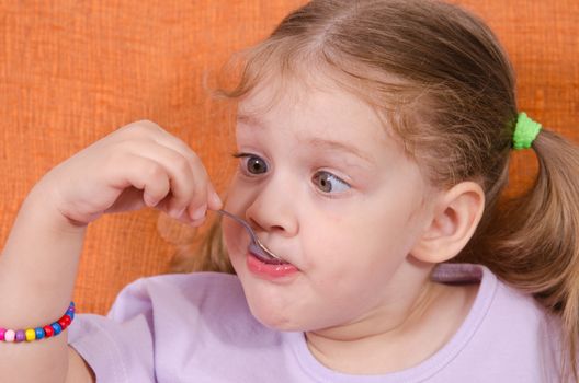 Three year old girl with pleasure and humor eats honey spoon sitting on the couch