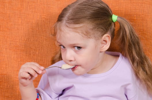 Three year old girl with pleasure and humor eats honey spoon sitting on the couch