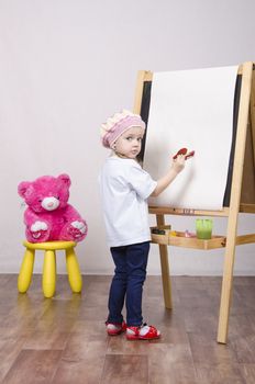 Three-year-old girl playing in the artist. Girl draws on the easel paints bear sitting on a chair