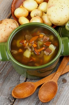 Delicious Vegetarian Soup with Chanterelle Mushrooms  in Green Pot with Raw Potato, Onion, Marinated Mushrooms and Wooden Spoons on closeup Rustic Wooden background. Top View