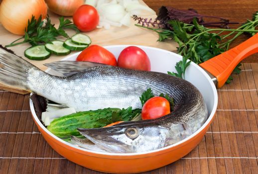 On a table on a chopping board there are a fish, cucumbers, tomatoes, onions, spices and parsley greens.