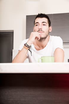 A young man sitting in the kitchen and drinking coffee.