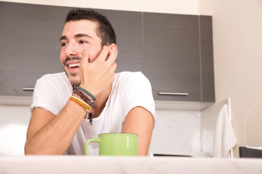 A young man sitting in the kitchen and drinking coffee.