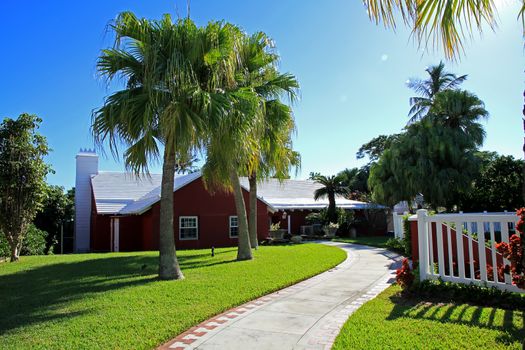 A single story, red walled, traditional Bermuda building with palm trees in the garden