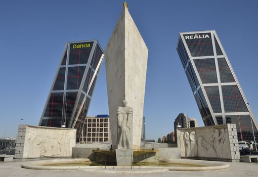 Monument to Calvo Sotelo, a Spanish politician, in Plaza Castilla, with the KIO towers. KIO Towers are twin office buildings in Madrid, Spain.  KIO Towers, are twin office buildings in Madrid, Spain. 
They were designed by the American architects Philip Johnson and John Burgee, commissioned by the Kuwait Investment Office (hence their initial name "Torres KIO" or "KIO Towers").  Each building is 115 metres tall with an inclination of 15 degrees, making them the first inclined skyscrapers in the world. 
