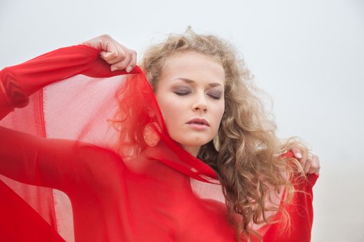 Beautiful woman with closed eyes in red fabric posing on sea beach in foggy day