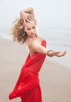 Beautiful nude woman in red fabric posing on sea beach in foggy day