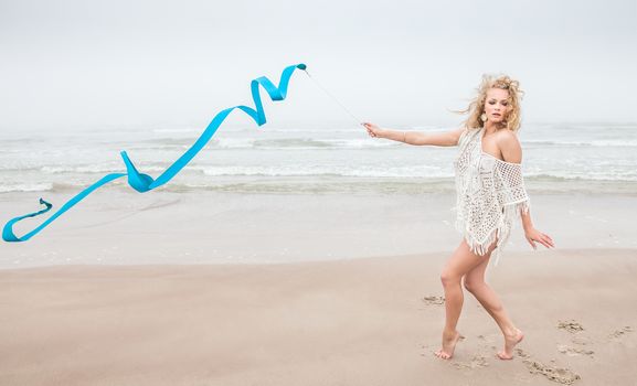 Young beautiful gymnast woman dance with ribbon on the beach at foggy day