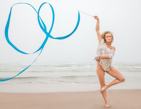 Young beautiful gymnast woman dance with ribbon on the beach at foggy day