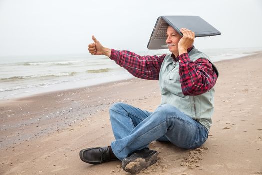 All OK. Old man standing on the beach with a laptop and a raised thumb up.