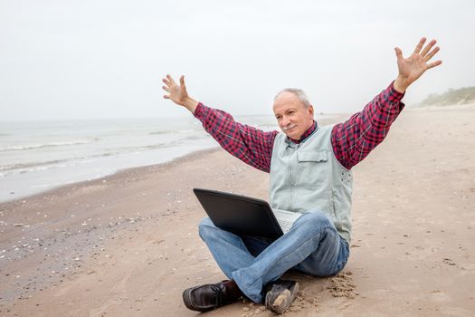 Happy old man sitting on the beach with a laptop and raising his hands