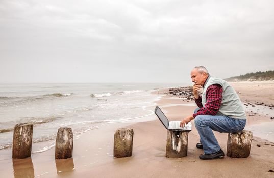 Thoughtful elderly man with the laptop sits on a sea background