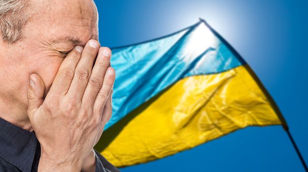 The war in Ukraine. Elderly man covered his face against of Ukrainian flag with bloody handprints
