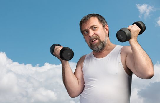 Elderly man exercising with dumbbells on blue sky with clouds background