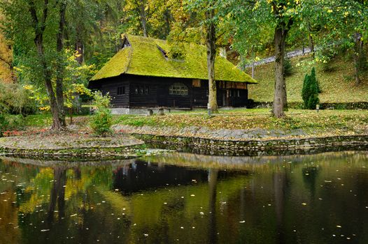 black wooden house with green moss on roof in autumn park with lake