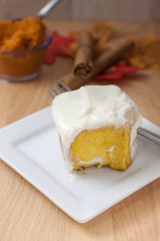 Homemade pumpkin cinnamon roll on a white plate with pumpkin, cinnamon, and fall leaves on a wooden table.