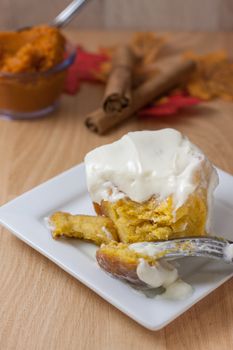 Homemade pumpkin cinnamon roll on a white plate with pumpkin, cinnamon, and fall leaves on a wooden table.