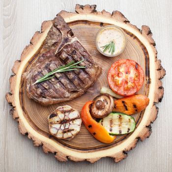 Portion of BBQ t-bone steak  served  on wooden board with  rosemary, mustard sauce  and grilled vegetables : tomato, carrot, paprika, garlic,  champignon,  zucchini