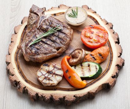 Portion of BBQ t-bone steak  served  on wooden board with  rosemary, mustard sauce  and grilled vegetables : tomato, carrot, paprika, garlic,  champignon,  zucchini