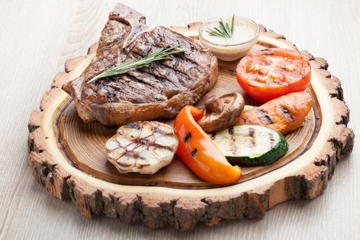 Portion of BBQ t-bone steak  served  on wooden board with  rosemary, mustard sauce  and grilled vegetables : tomato, carrot, paprika, garlic,  champignon,  zucchini