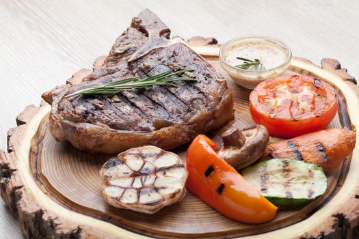 Portion of BBQ t-bone steak  served  on wooden board with  rosemary, mustard sauce  and grilled vegetables : tomato, carrot, paprika, garlic,  champignon,  zucchini