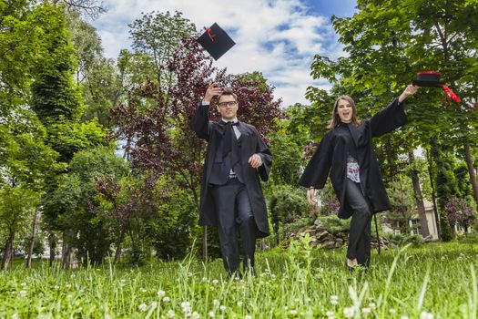 Happy couple in the graduation day running in a park and throwing  their hats up.